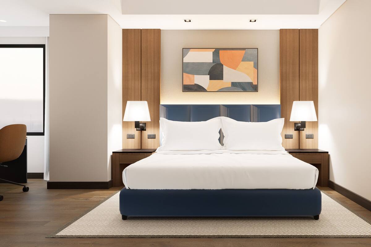 A hotel room with a large bed, two bedside lamps, a modern abstract painting, and a chair.
