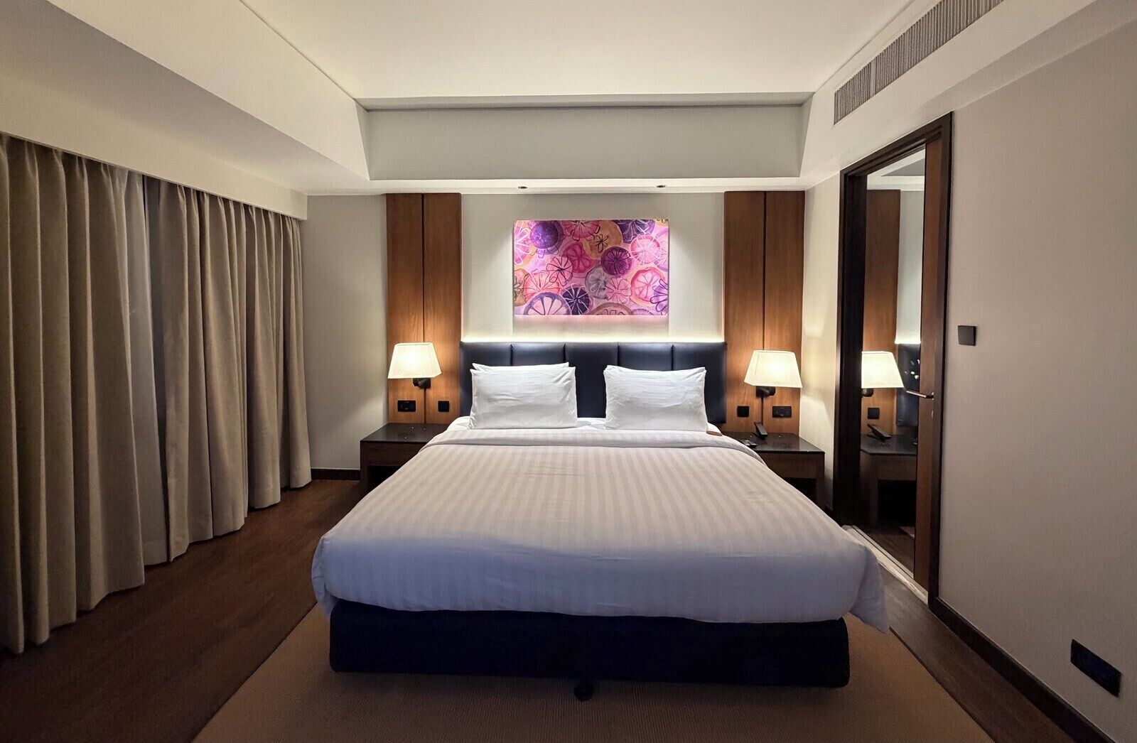 A hotel room with a large bed, two bedside tables with lamps, a large mirror, and a window with curtains.