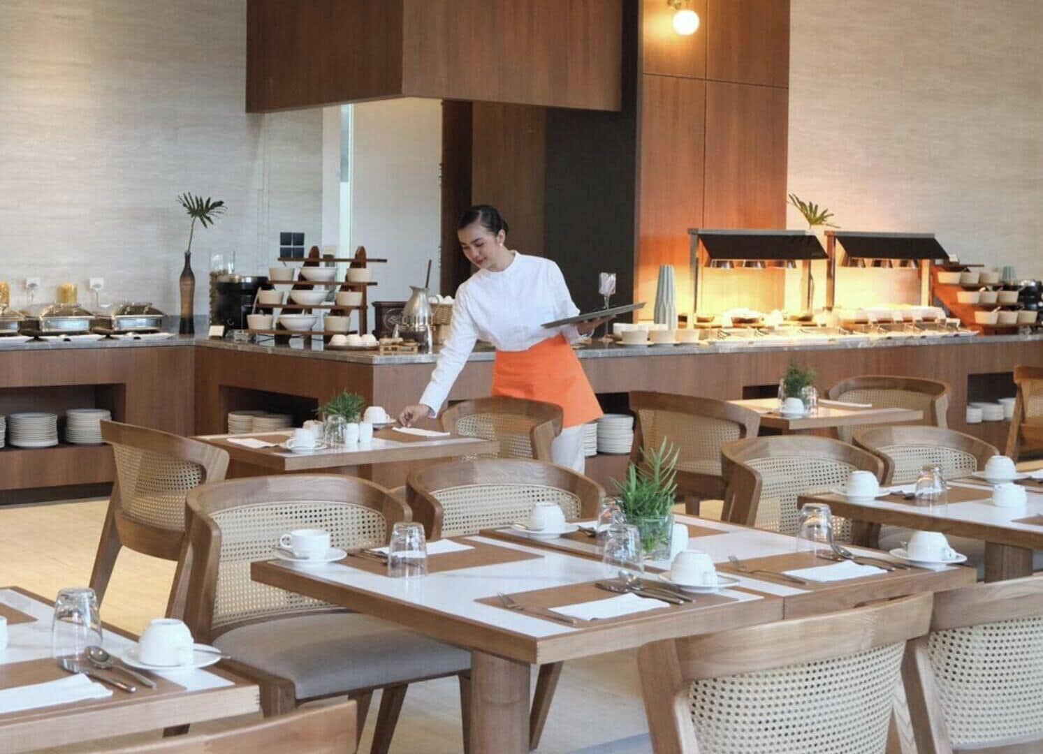 A restaurant with wooden tables and chairs, a buffet area with various dishes, and a person serving food.