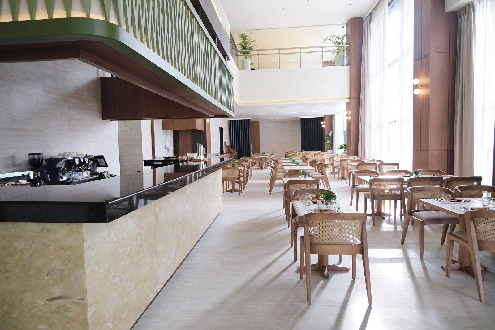 Un comedor amplio con varias mesas y sillas, una barra larga con una cafetera, y ventanas grandes que dejan entrar luz natural.