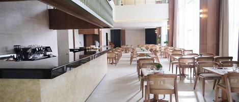 A spacious dining area with multiple tables and chairs, a long counter with a coffee machine, and large windows allowing natural light.