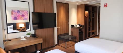 A hotel room with a bed, a desk with a chair, a TV, a wardrobe, and a small seating area.
