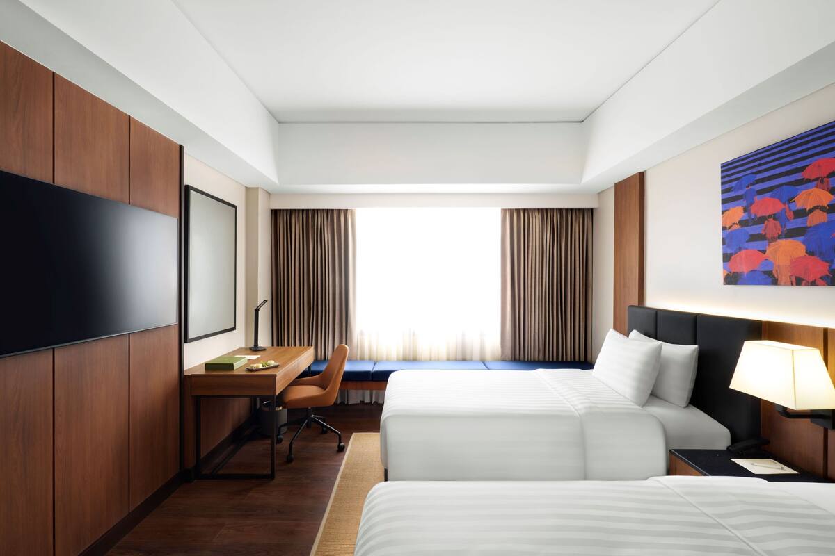 A hotel room with two beds, a desk, a chair, a TV, and a painting on the wall.