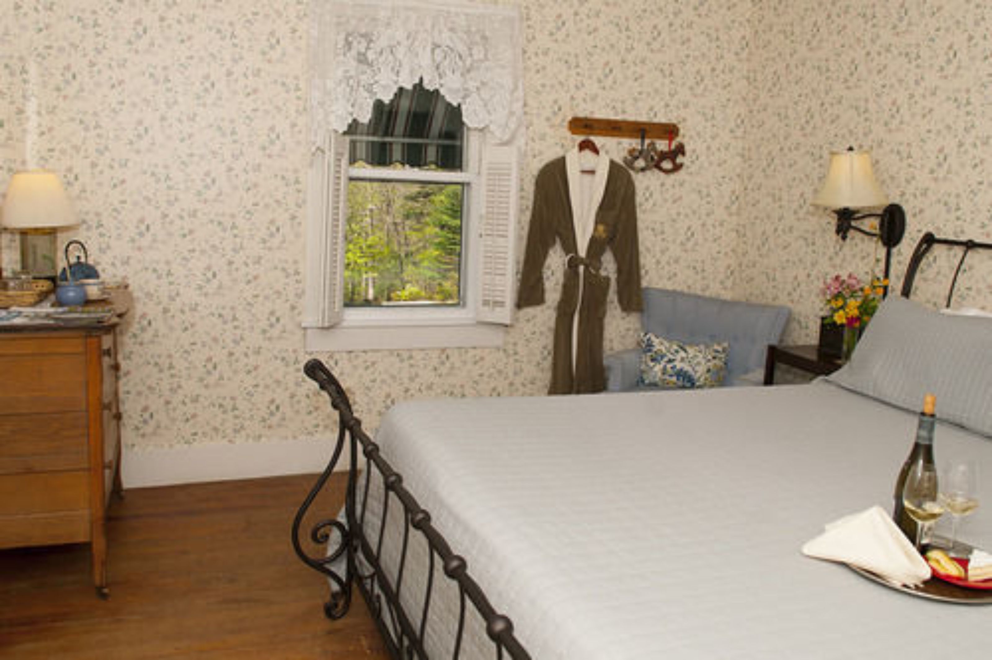 Quarto com cama, cômoda, janela com vista, roupão pendurado na parede e bandeja com garrafa e copos.