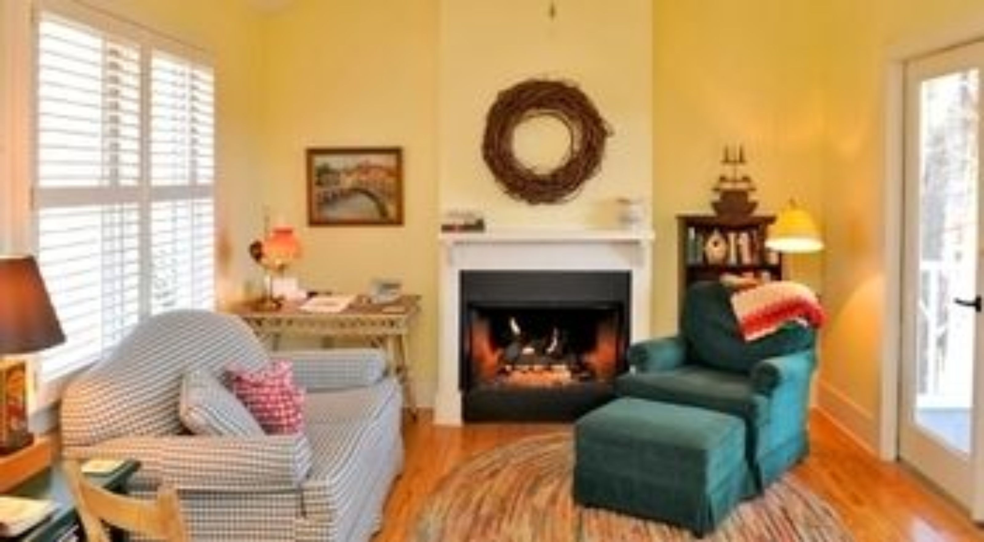 Un salon chaleureux avec une cheminée, un fauteuil en osier, un fauteuil de couleur turquoise, un plancher en bois et une fenêtre avec des stores.