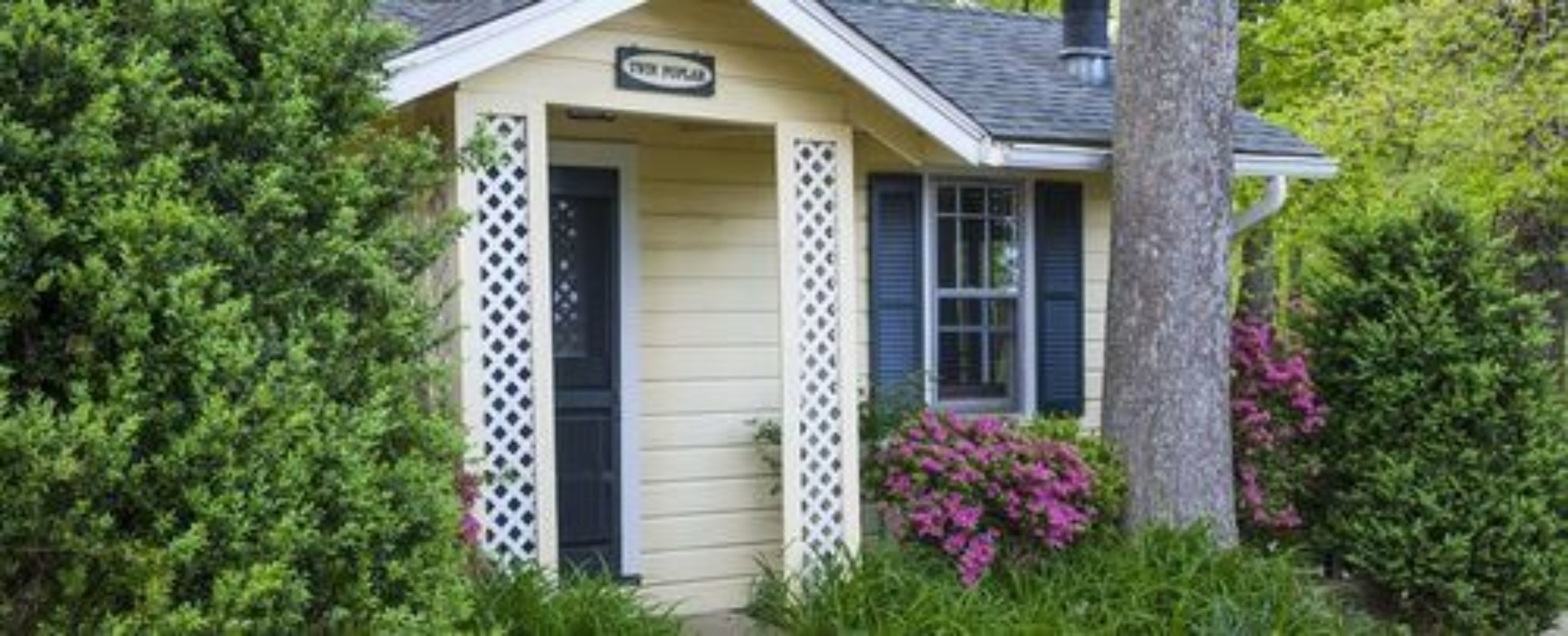 A small, single-story building with a sign that reads "The Pub." It features a white lattice door and window shutters, surrounded by lush greenery and flowering plants.