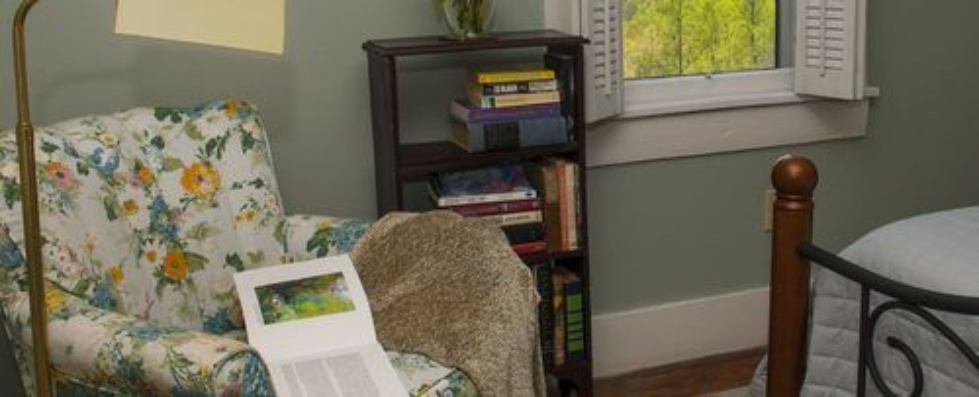 Una habitación con un sillón de estampado floral, una estantería con libros y una ventana con persianas.