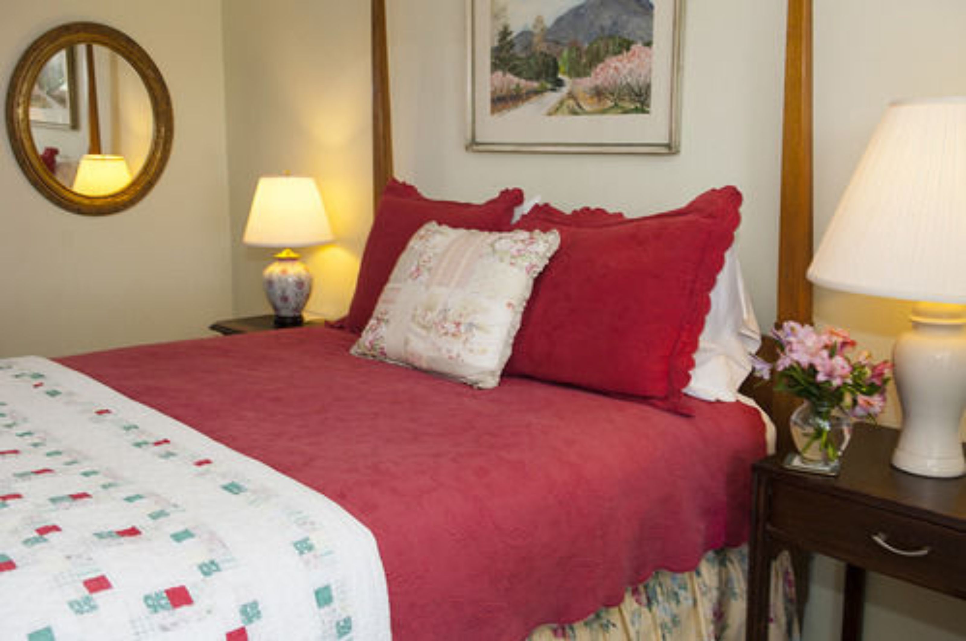 A neatly made bed with a red blanket and pillows, a bedside table with a lamp, and a framed picture on the wall.