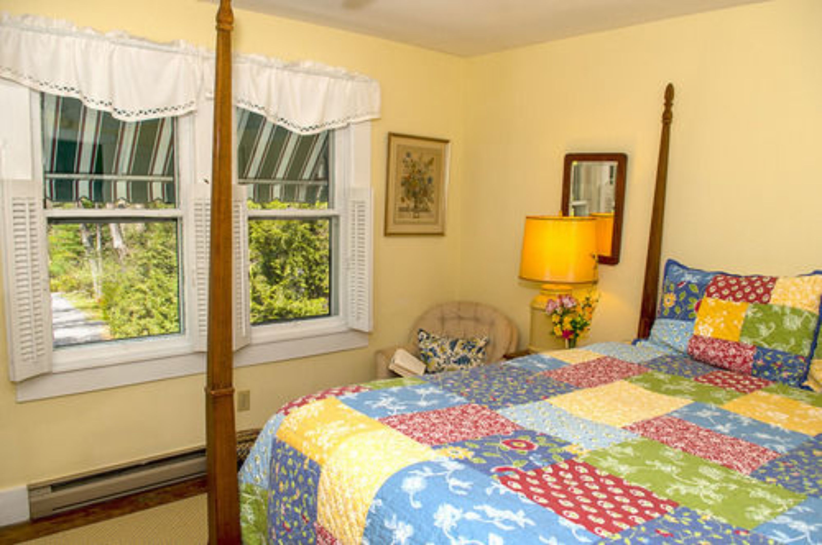 Un dormitorio con cama de dosel, una ventana con contraventanas, una lámpara de noche y un arreglo floral.