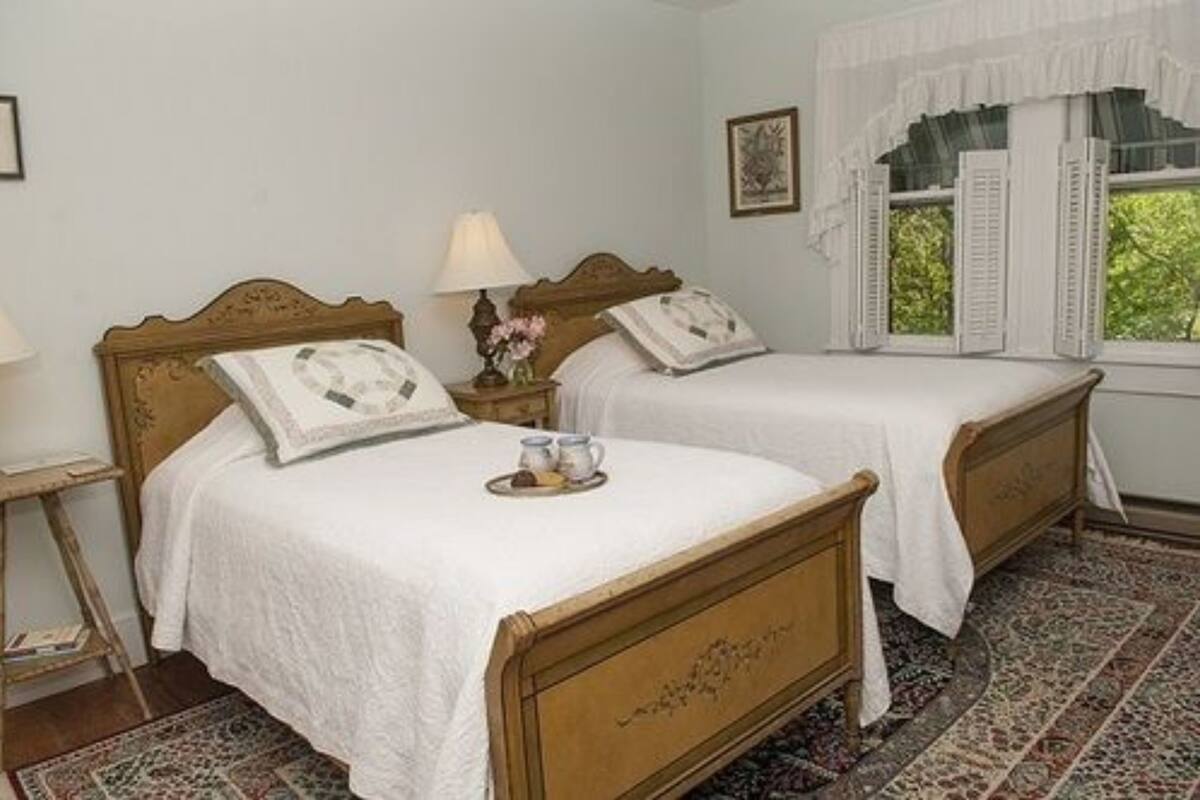 A bedroom with two wooden beds, white bedding, a nightstand with a lamp, a small table with a tray, and a window with shutters.