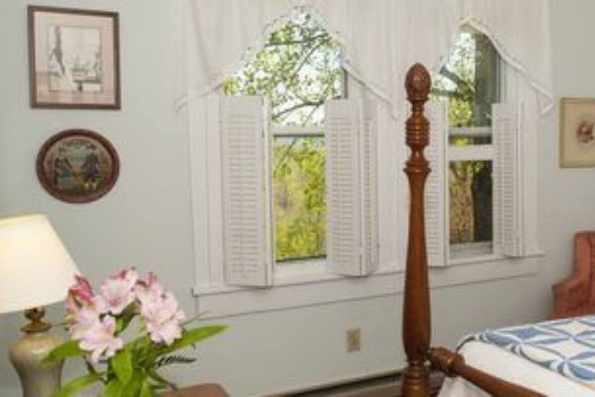 A bedroom with a four-poster bed, a window with shutters, a bedside lamp, and a floral bedspread.