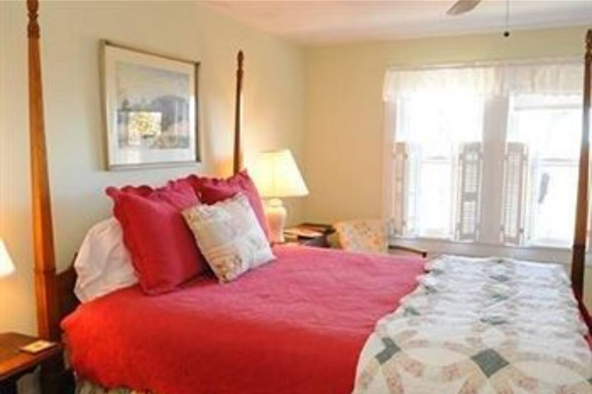 A bedroom with a four-poster bed, a nightstand, a lamp, and a window with curtains.