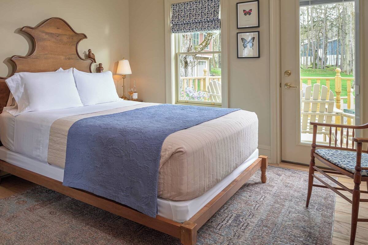 A neatly made bed with a wooden headboard, two bedside tables with lamps, a chair, and a view of a deck through a door.
