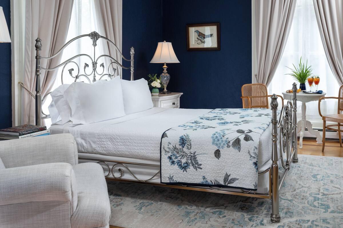 A neatly arranged bedroom with a bed, bedside table, lamp, chair, and a small table with drinks.