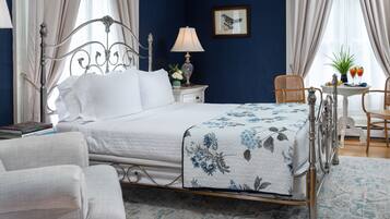 A neatly arranged bedroom with a bed, bedside table, lamp, chair, and a small table with drinks.