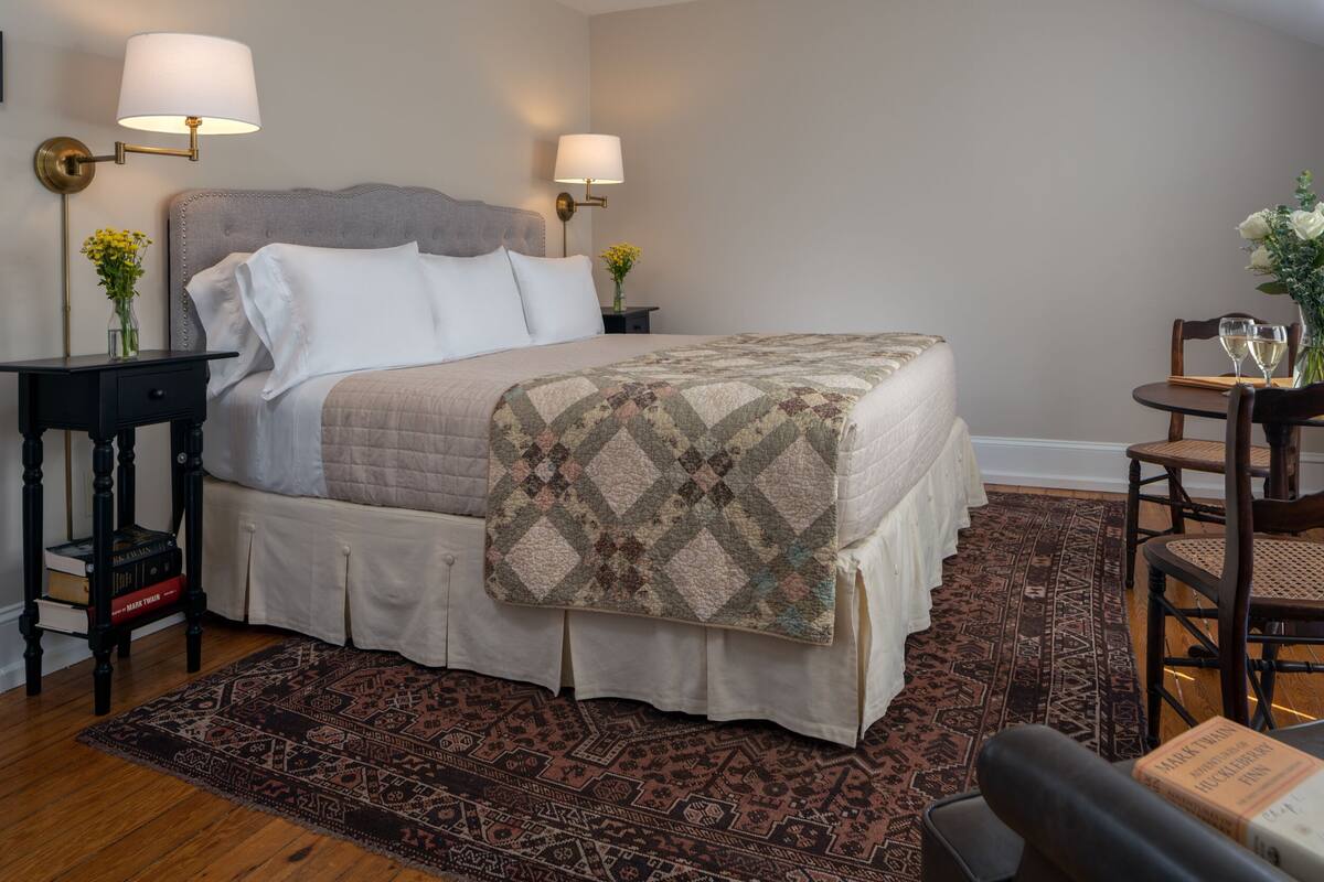 A neatly arranged bedroom with a large bed, two bedside tables with lamps, a small round table with chairs, and a patterned rug.