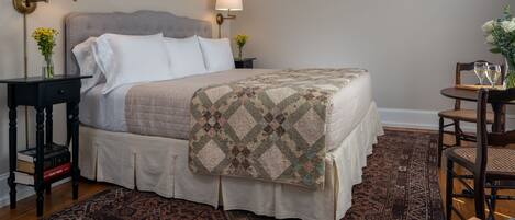 A neatly arranged bedroom with a large bed, two bedside tables with lamps, a small round table with chairs, and a patterned rug.