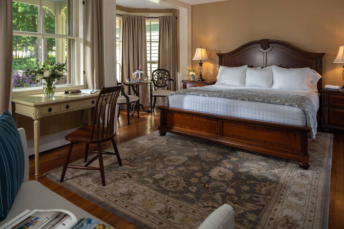 A hotel room with a large bed, a desk with a chair, a small table, and a window with curtains.