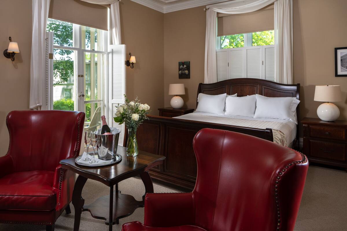A hotel room with a large bed, two red leather chairs, a wooden nightstand with lamps, and a window with curtains.