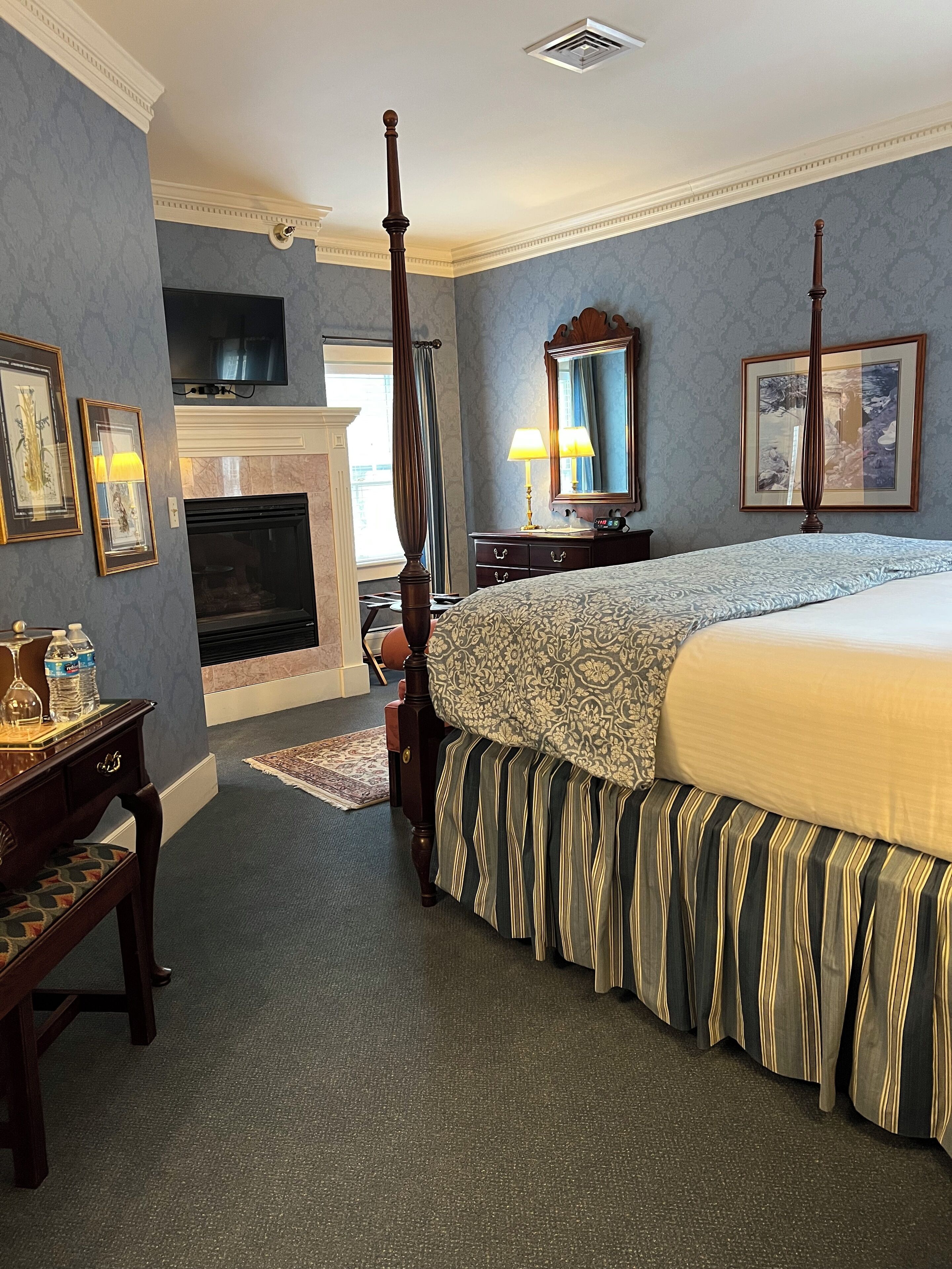 A bedroom with a four-poster bed, a fireplace, a television mounted on the wall, and framed artwork on the walls.