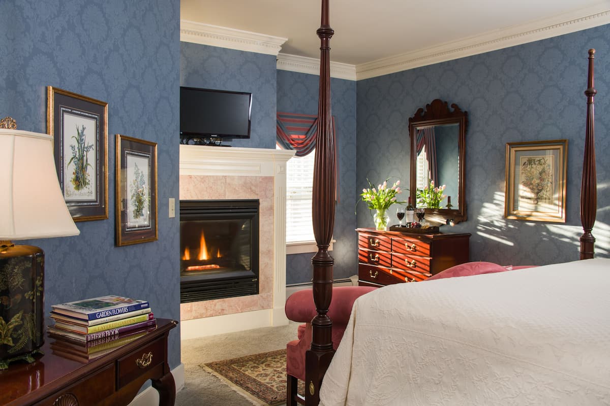 A bedroom with a four-poster bed, a fireplace, a television, and framed artwork on the walls.