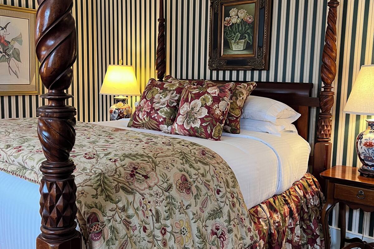 A four-poster bed with a floral bedspread, striped wallpaper, and a framed picture on the wall.
