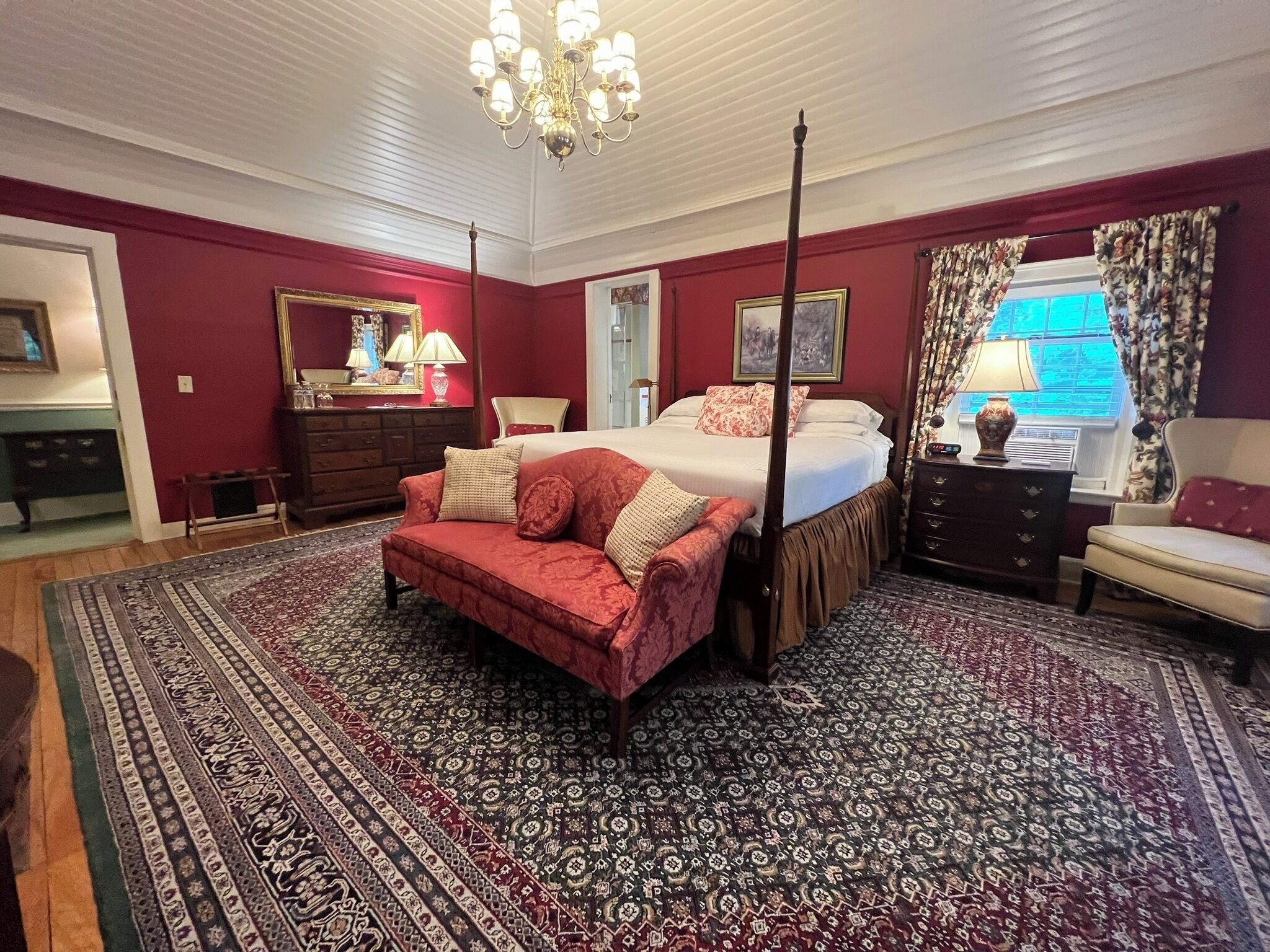 A four-poster bed with a canopy, a red sofa, and a patterned rug in a room with red walls and a chandelier.
