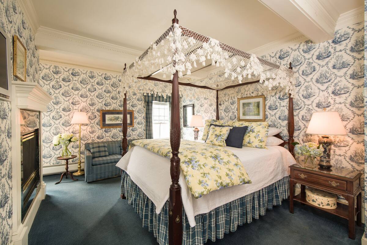 A four-poster bed with a canopy, a sofa, a bedside table with a lamp, and a small table with flowers.