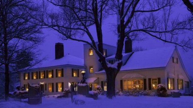 Une maison à deux étages, dont le toit et la cour sont recouverts de neige, avec de nombreuses fenêtres illuminées et une cheminée bien visible.