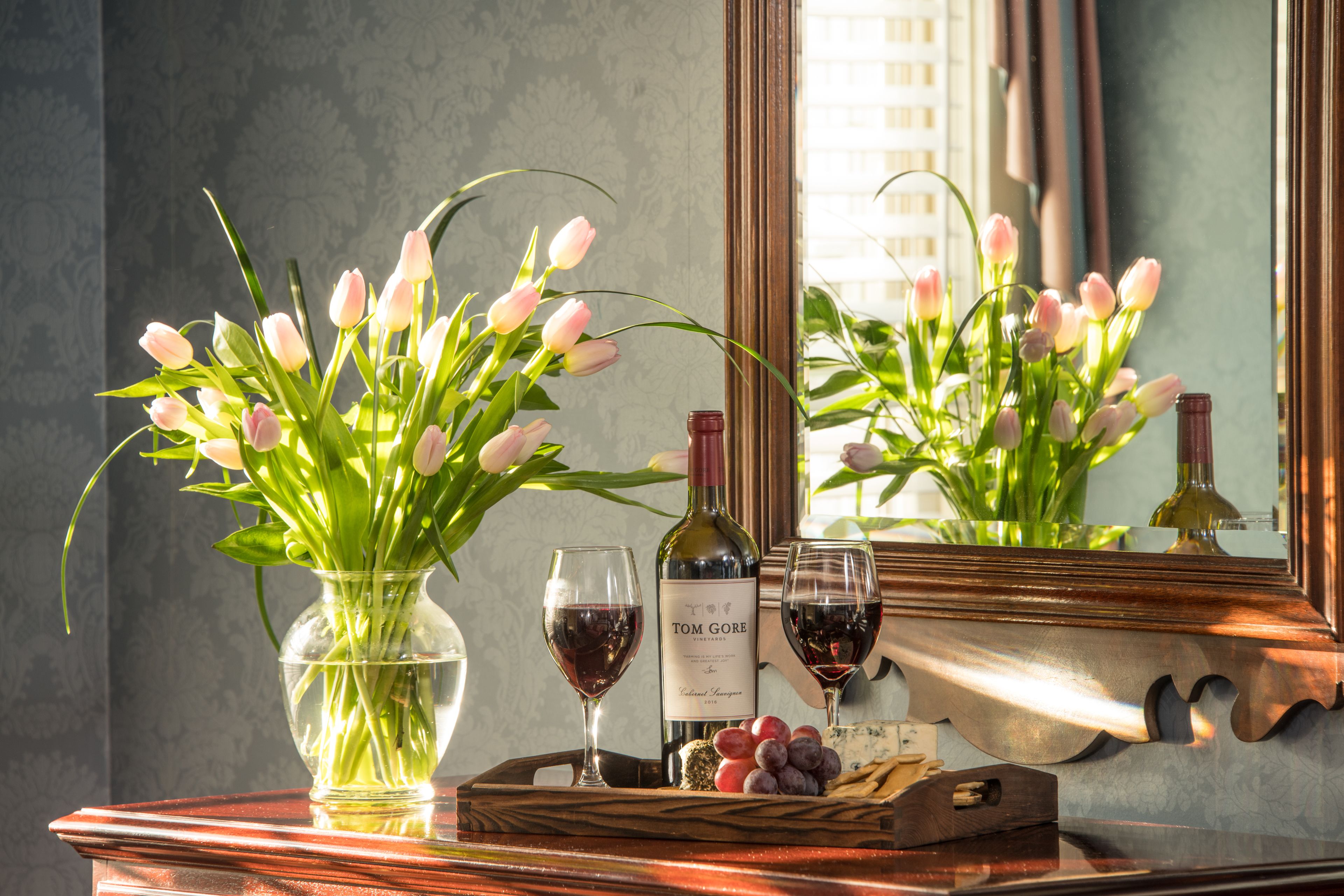 A table with a vase of pink tulips, two wine glasses, a bottle of wine, and a fruit tray.