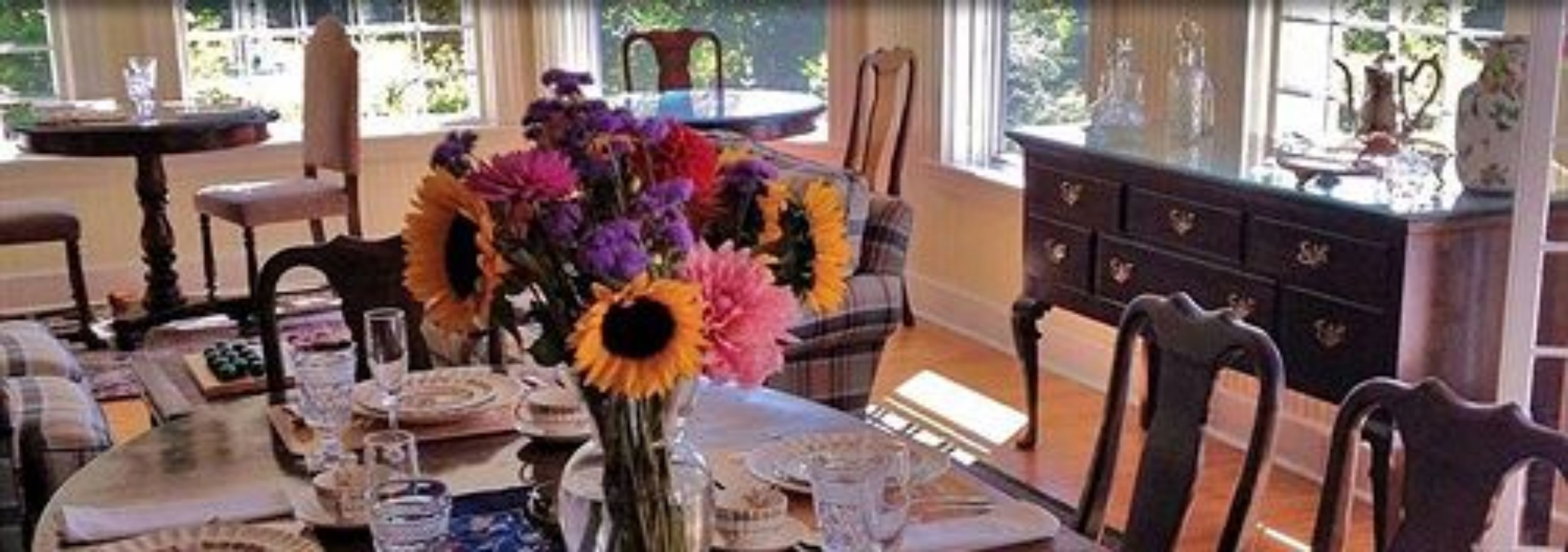 A dining area with a table set for a meal, a vase with flowers, and a view of the outdoors through large windows.