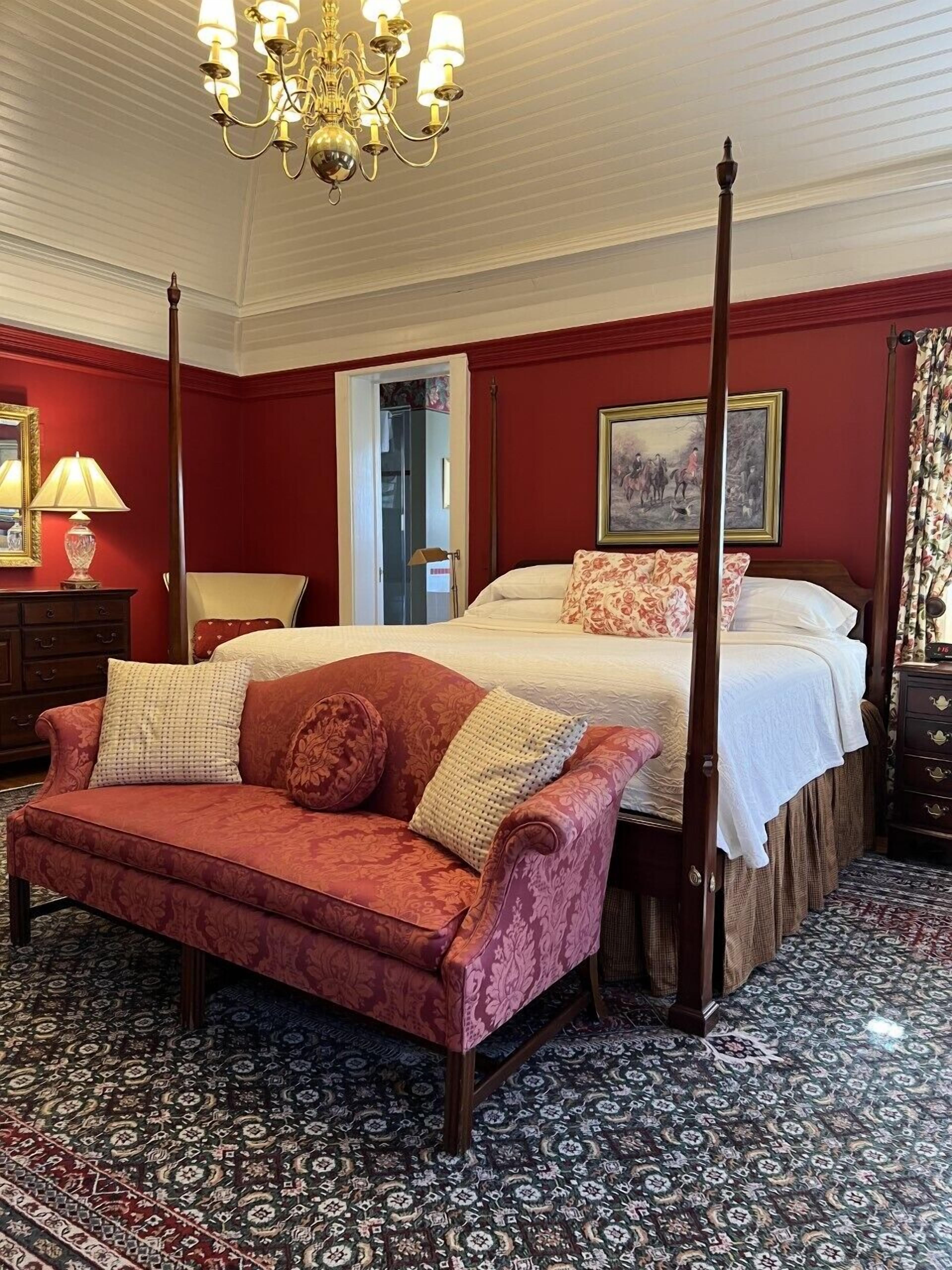 A four-poster bed with a white bedspread and red pillows, a red sofa with patterned cushions, and a chandelier.