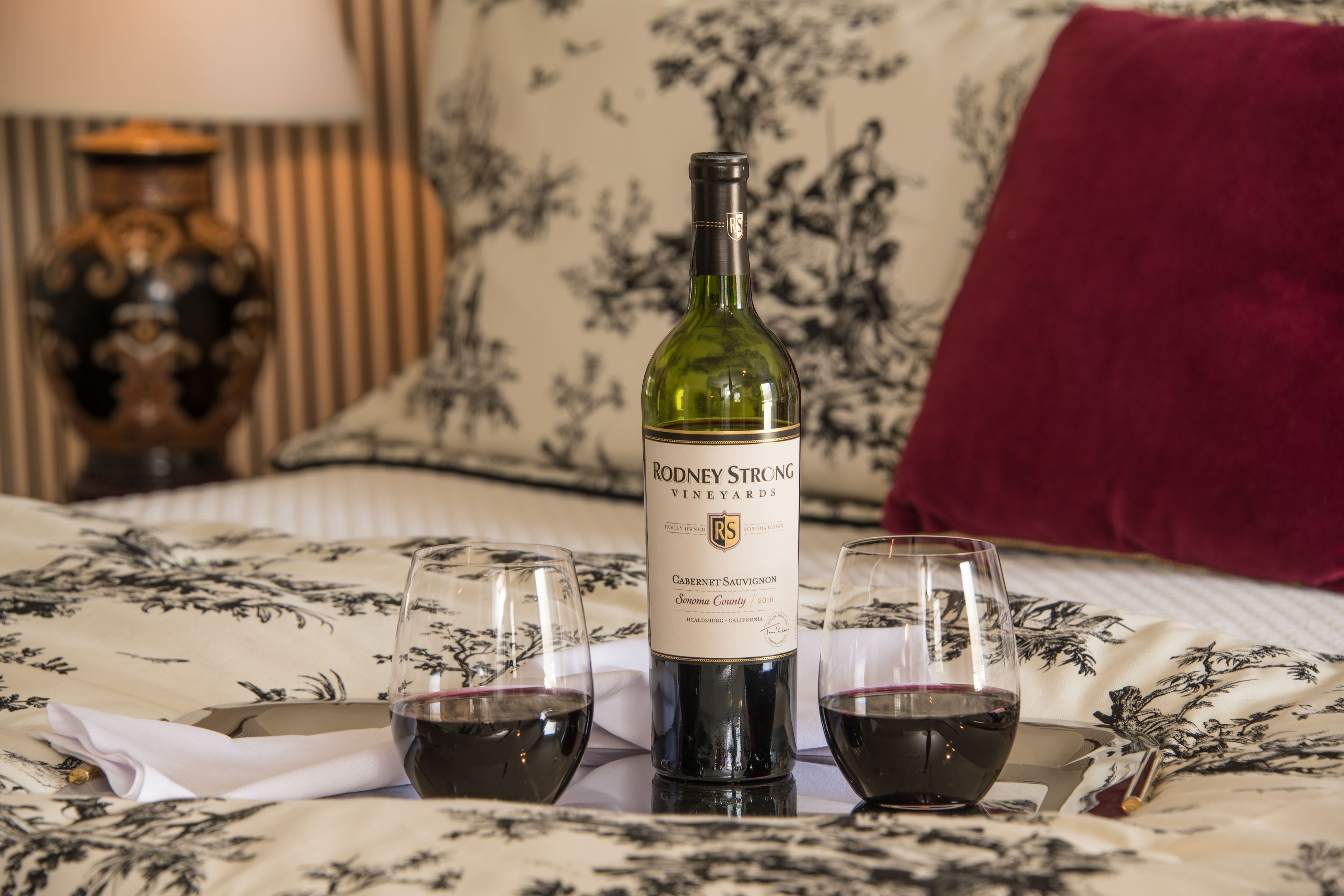 A bottle of Rodney Strong Vineyards Cabernet Sauvignon and two glasses of wine on a bed with a floral bedspread.