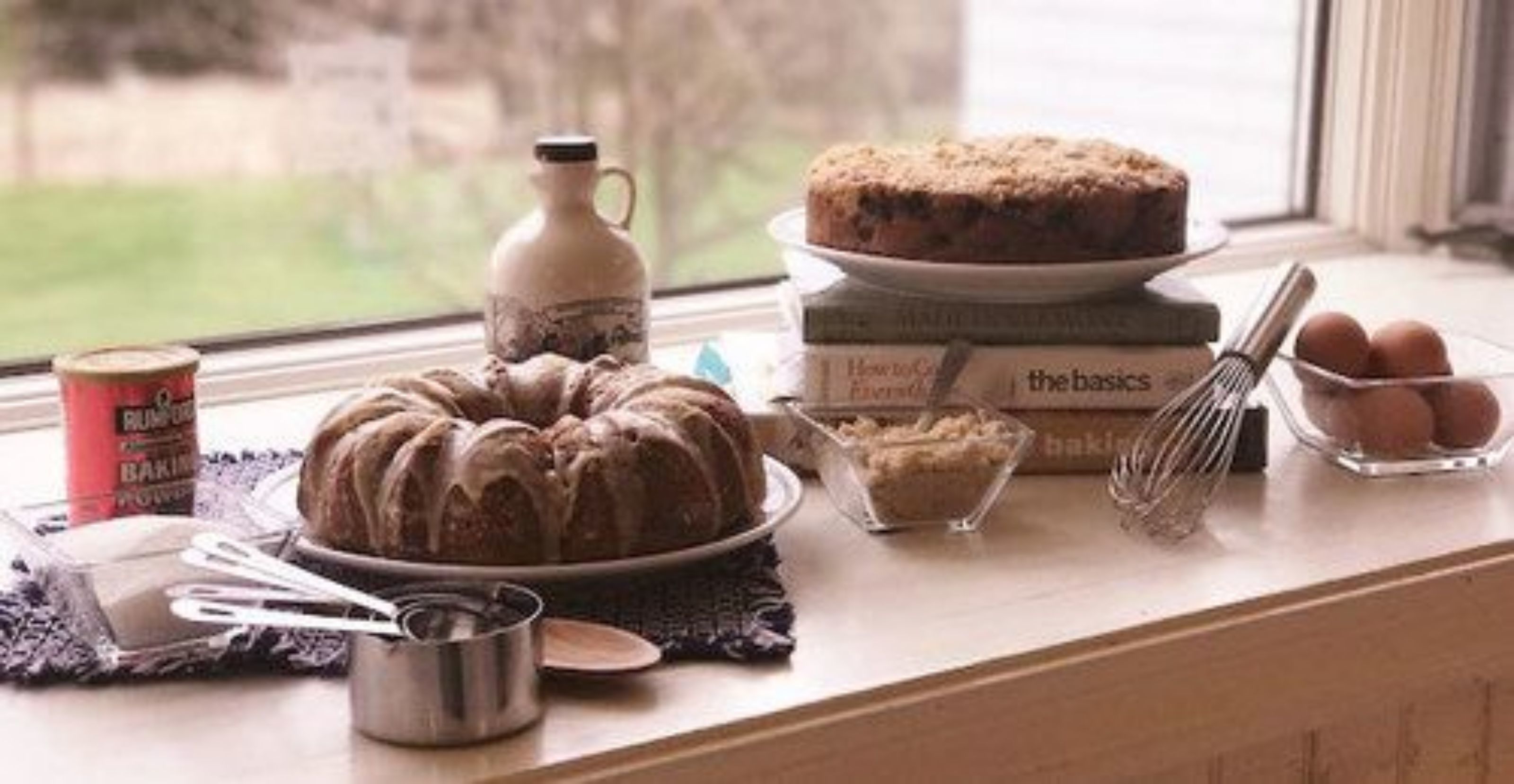 A kitchen counter with a bundt cake, a whisk, a bowl of flour, a bottle of baking powder, and a window with a view of greenery.