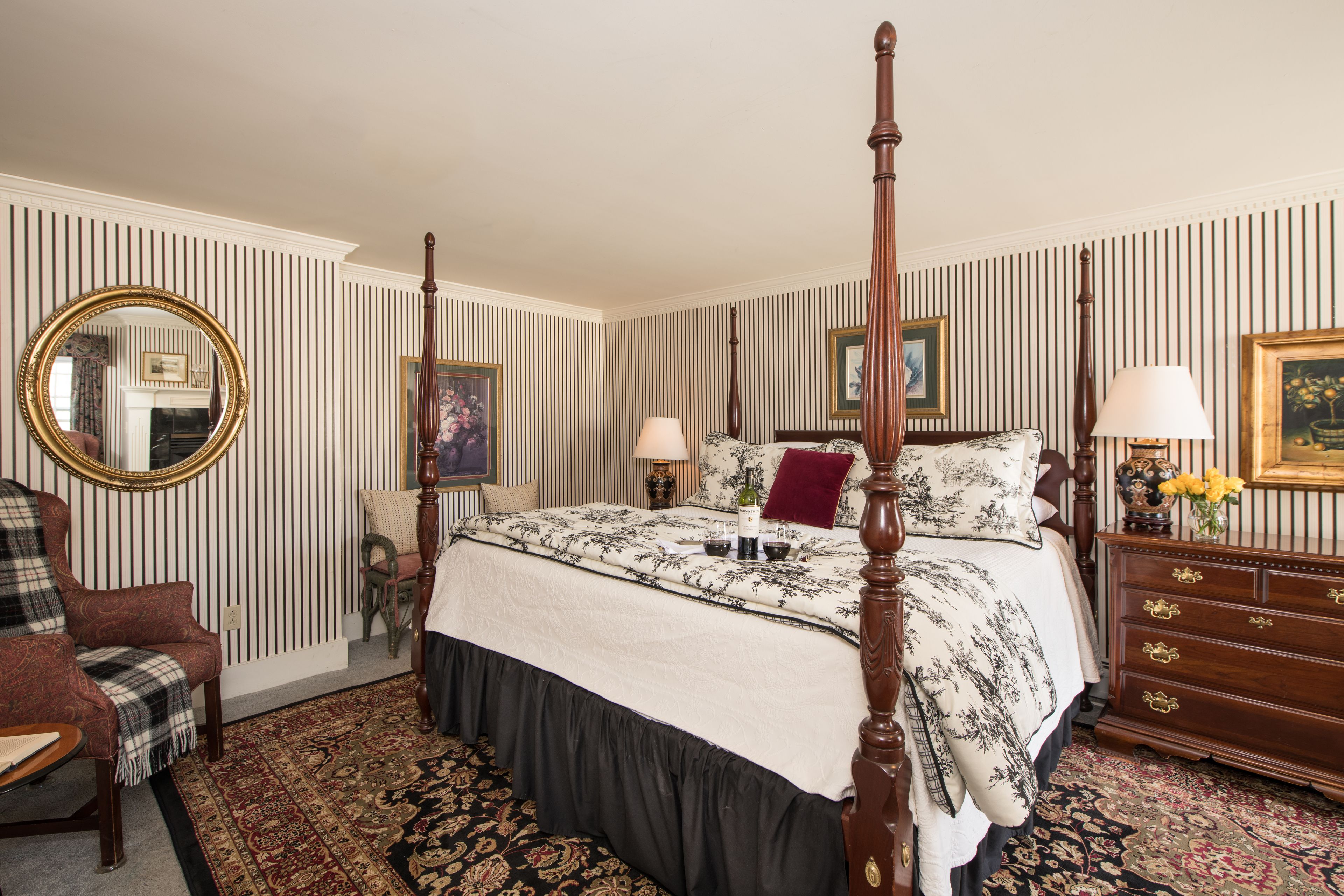 A four-poster bed with a floral bedspread, a wooden dresser with drawers, a round mirror, and a plaid armchair.