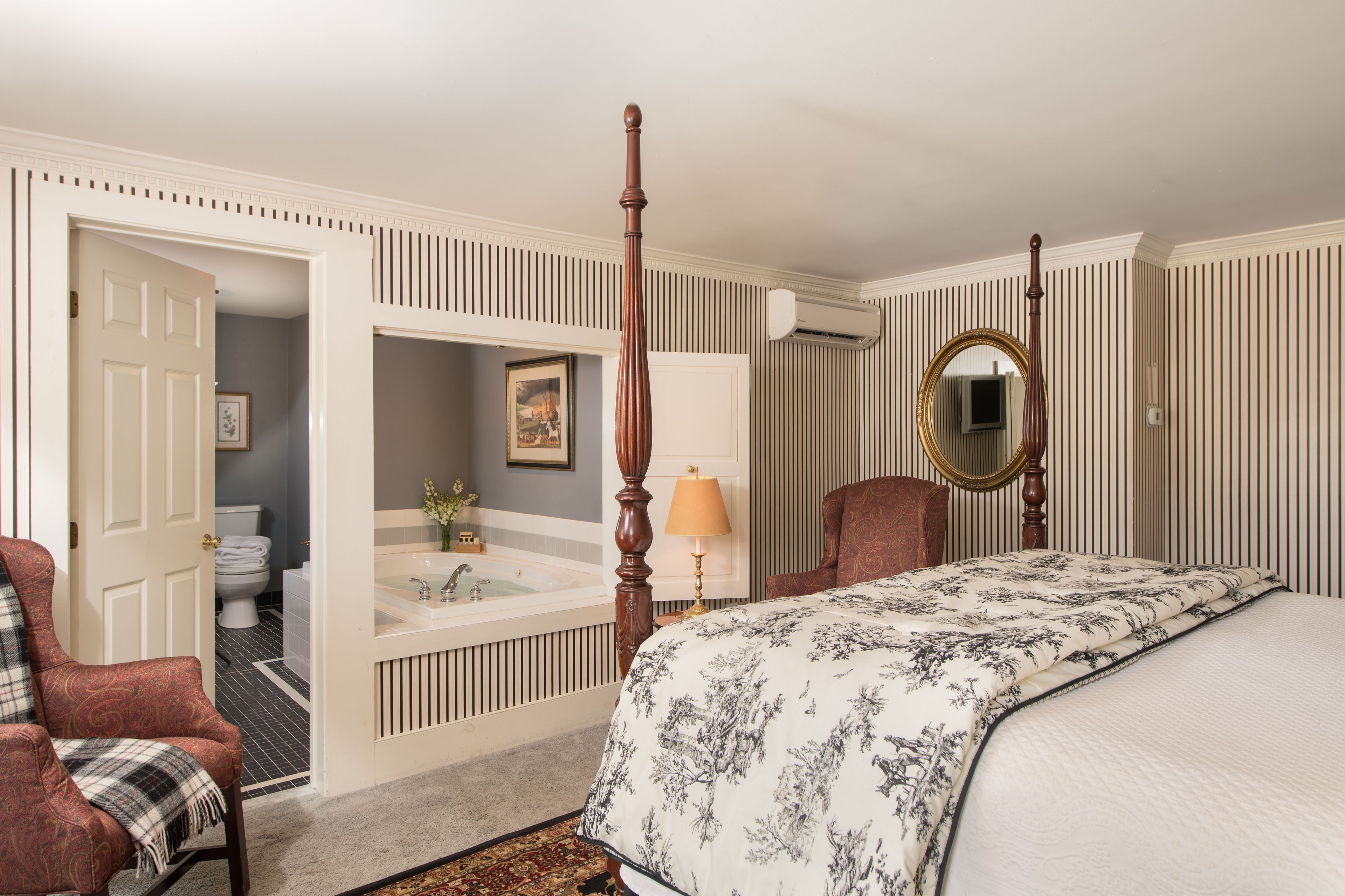 Un dormitorio con cama con dosel, una zona de estar con una silla, un baño con bañera y un espejo grande.