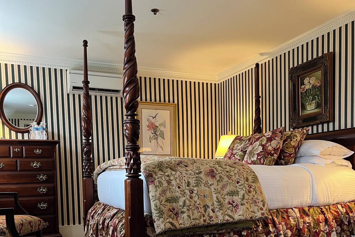 A bedroom with a four-poster bed, a dresser, a mirror, and striped wallpaper.