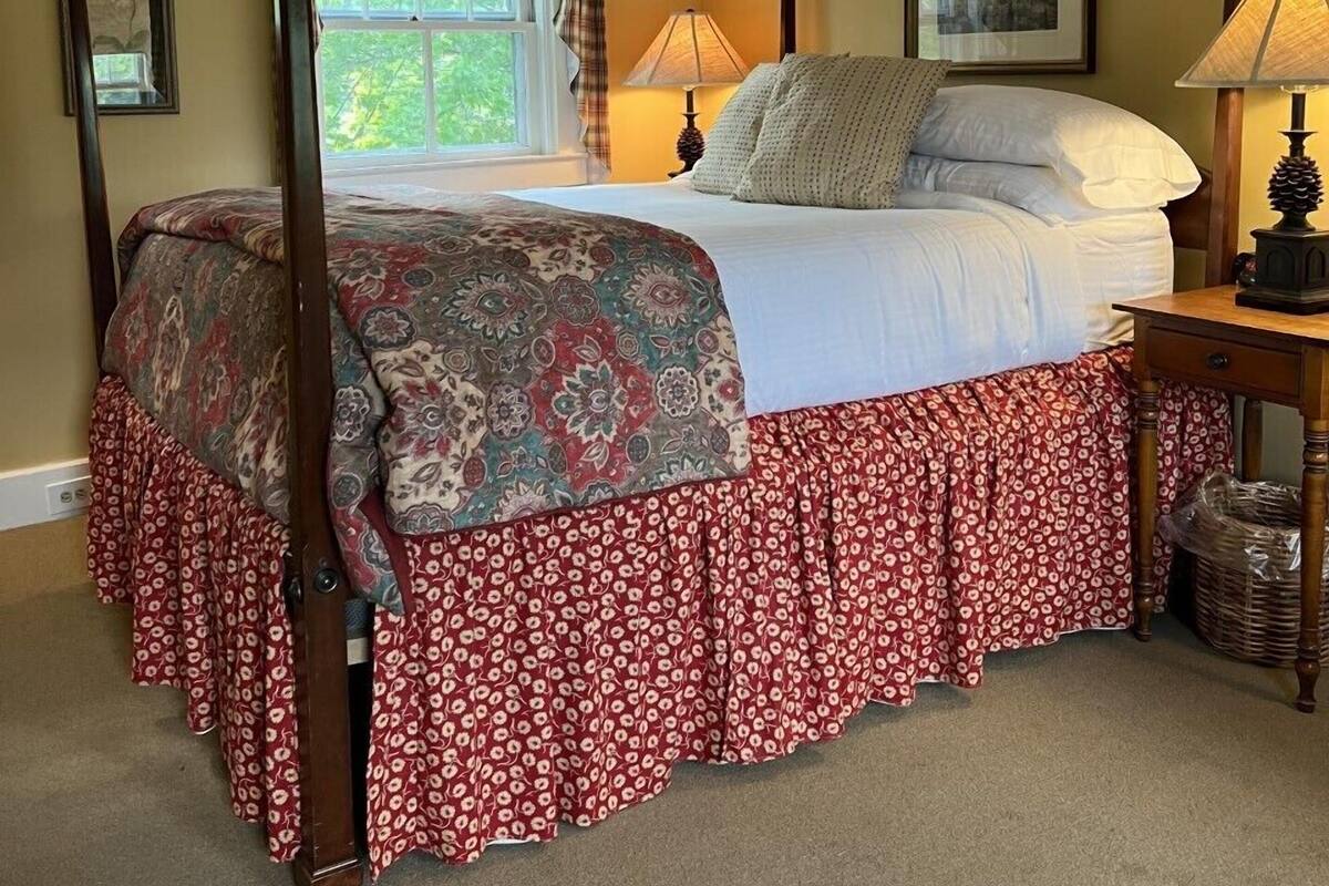 A four-poster bed with a floral bedspread and red skirted bed skirt, a window with curtains, a bedside table with a lamp, and a framed picture on the wall.