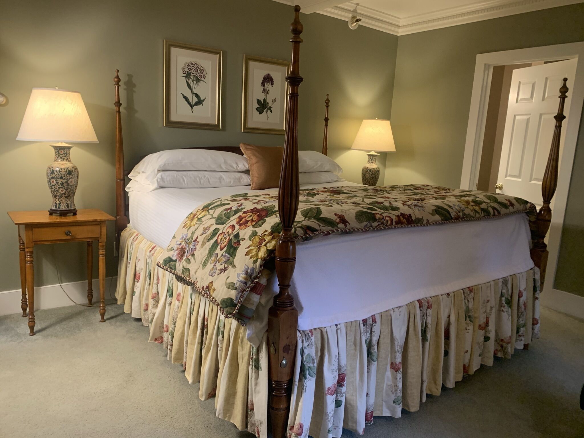 A four-poster bed with floral bedding, two bedside lamps, and framed botanical prints on the wall.