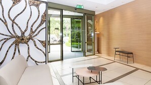 A modern lobby with a glass door entrance, a seating area with a sofa and tables, and a decorative wall panel. - Athinais Hotel (Athens)