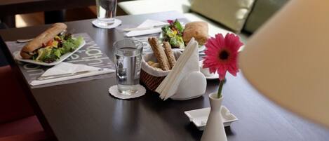 Una mesa de comedor con un sándwich, ensalada, pan y un vaso de agua. Hay un florero con una flor y una servilleta sobre la mesa.