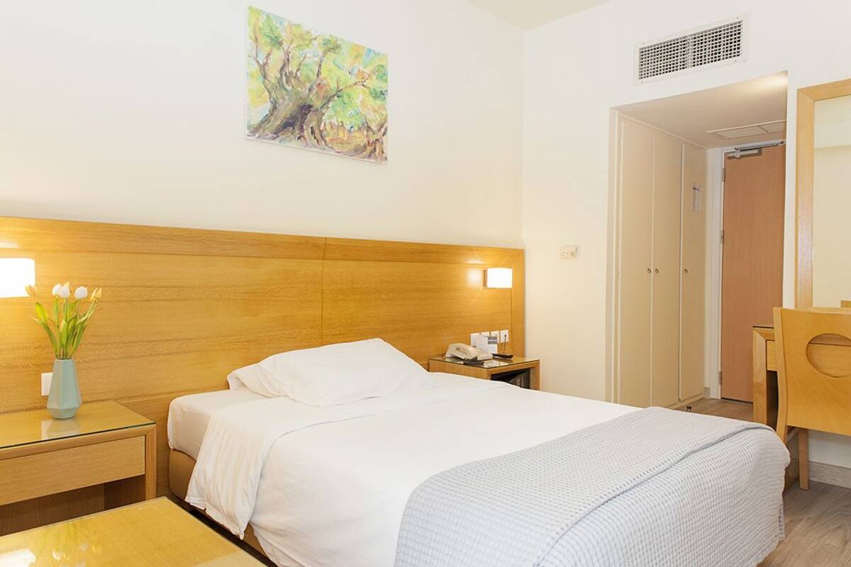 A hotel room with a wooden headboard, a bed with white linens, a nightstand with a vase of flowers, a desk with a telephone, and a painting on the wall.