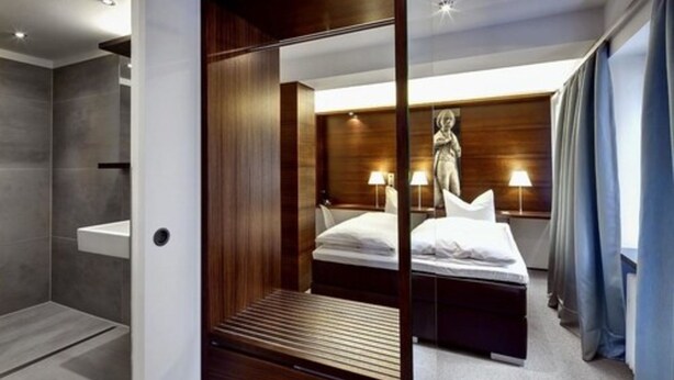 A modern hotel room with a bed, bedside lamps, a wooden headboard, and a shower area. - Stern am Rathaus (Cologne)