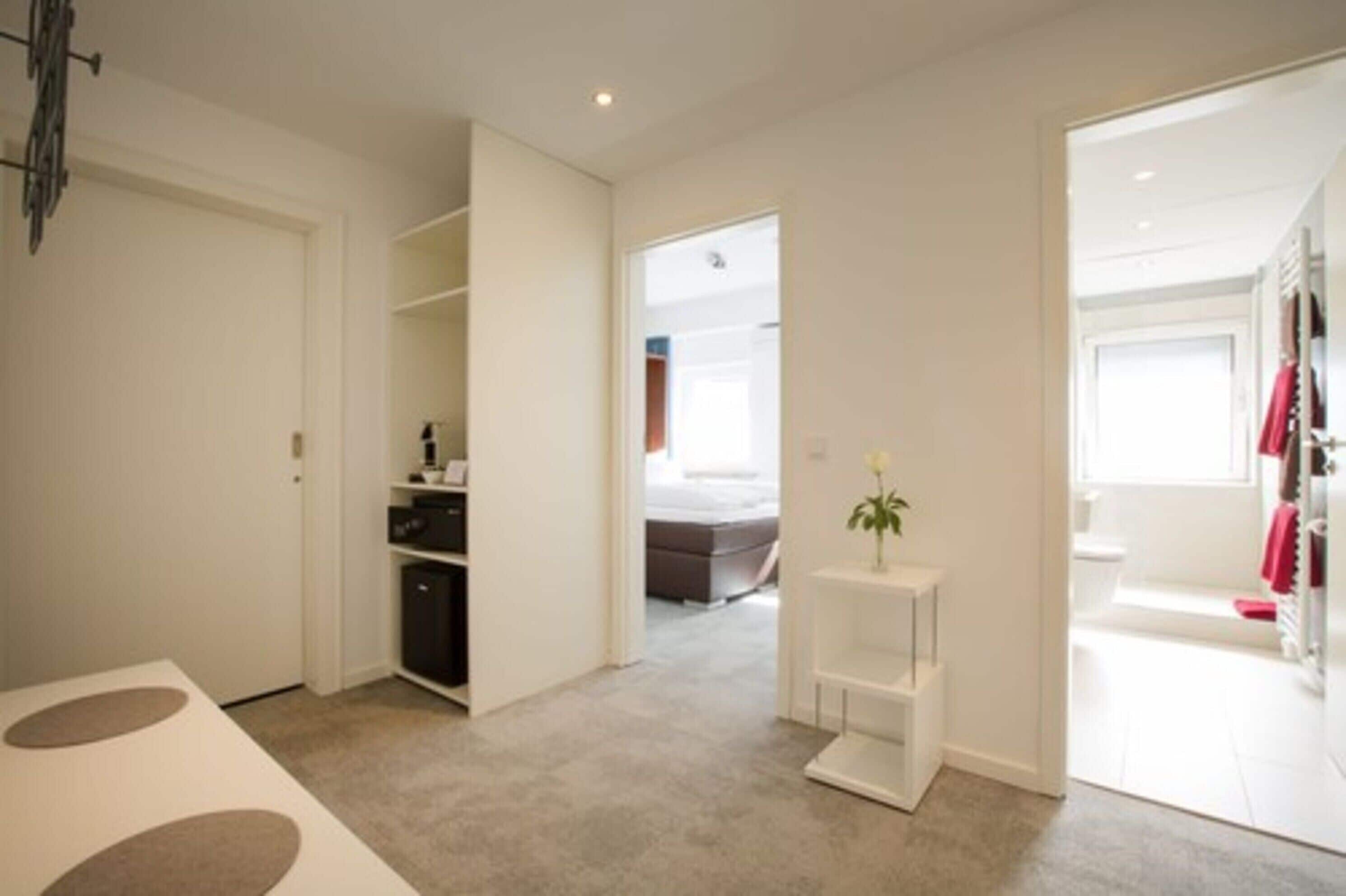 A modern, well-lit room with a neutral color palette, featuring a bed, a small table with a vase, and a closet with hanging clothes.