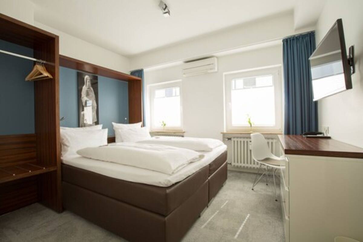 Quarto de hotel com cama de casal, escrivaninha com cadeira, guarda-roupa e televisão fixada na parede.