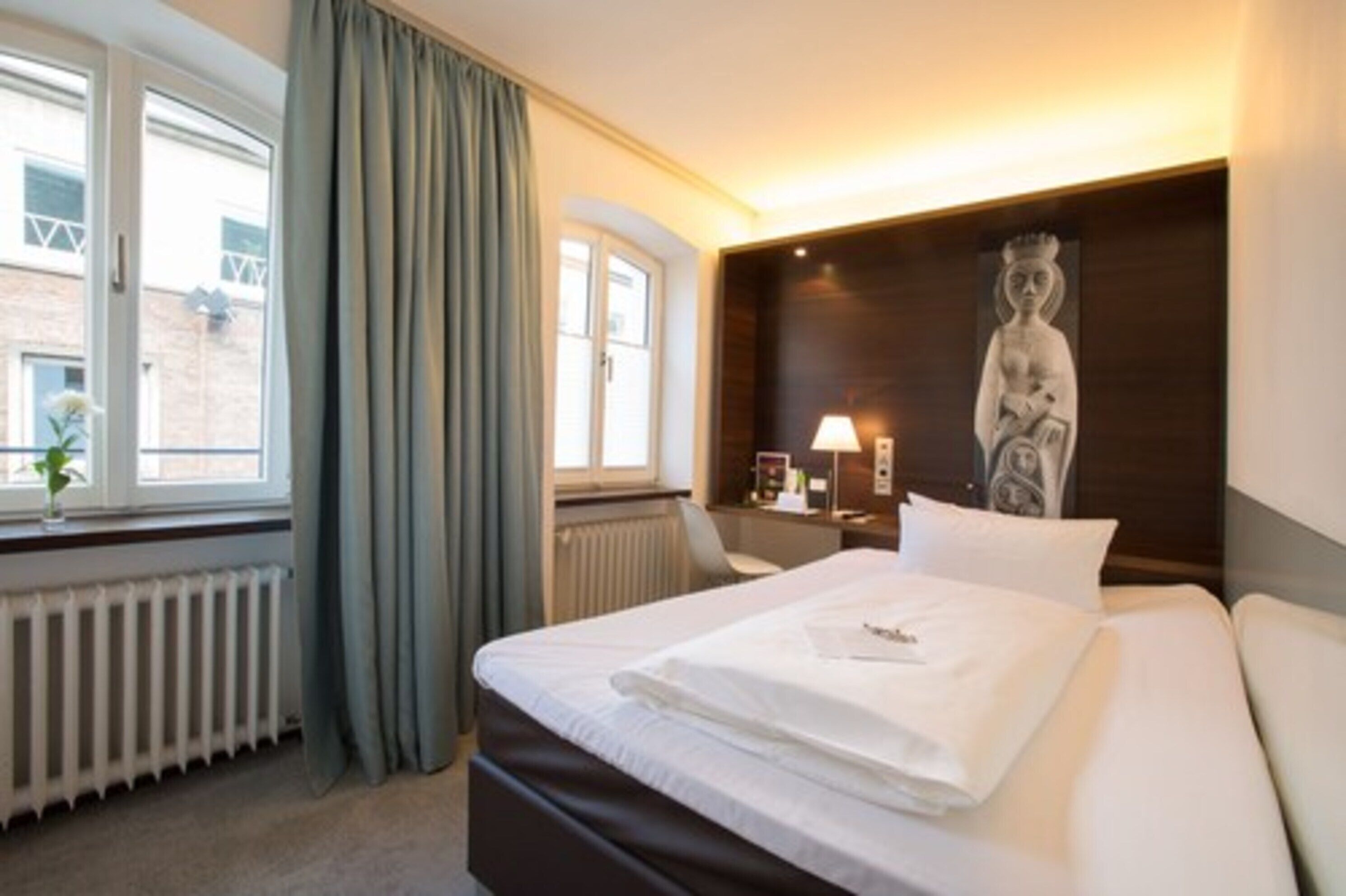 Una camera d'albergo con un letto, una finestra con tende, un termosifone, una scrivania con una lampada e una statua appesa al muro.
