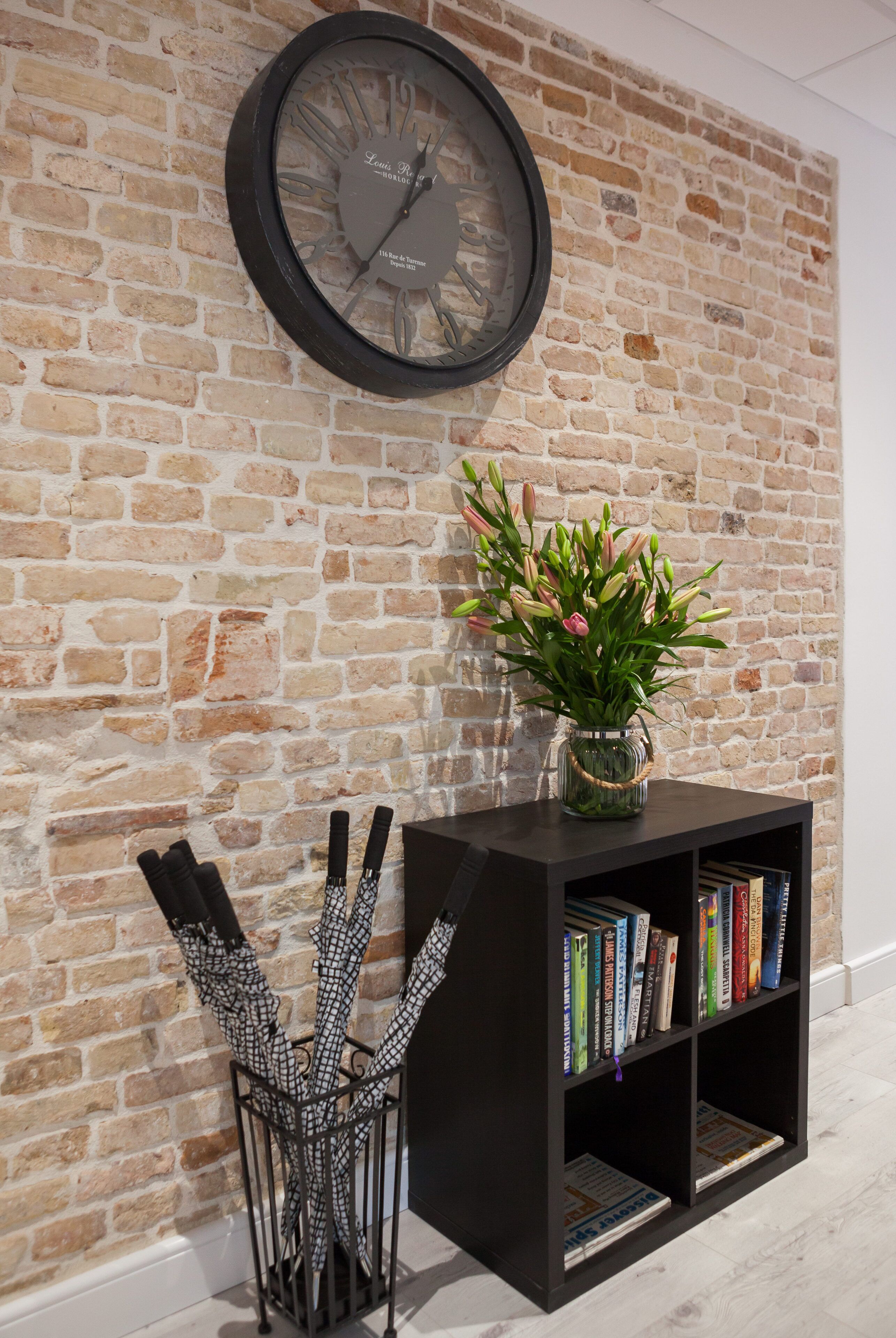 Un muro de ladrillo con un reloj redondo, una estantería llena de libros y un jarrón con flores.