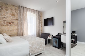 A modern hotel room with a brick wall, a large bed with a patterned bedspread, a desk with a computer, and a flat-screen TV mounted on the wall.