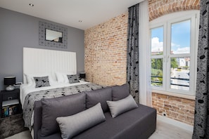 A bedroom with a large bed, a grey sofa, a brick wall, and a window with a view of buildings.