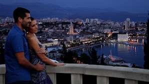 Un couple se tient sur un balcon, d’où l’on peut admirer une vue sur la ville, avec une rivière et des montagnes en arrière-plan.